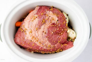 A slab of raw corned beef covered with mustard seeds is placed in a white slow cooker. Carrots and onions are partially visible beneath the beef, ready for slow cooking.