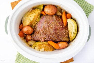 A slow cooker with corned beef, halved cabbage, whole red potatoes, and carrot pieces. The ingredients are seasoned with mustard seeds and spices. The cooker sits on a green and white checkered cloth.