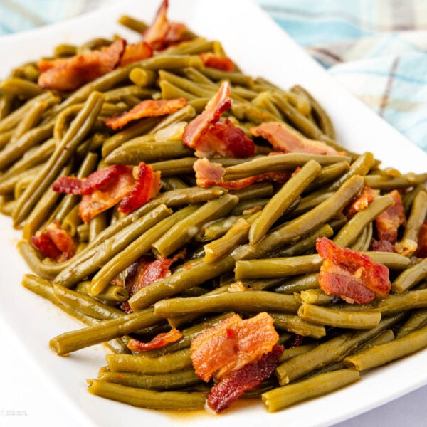 A rectangular white dish filled with cooked green beans mixed with crispy bacon pieces. The beans and bacon are evenly distributed, creating a rustic and appetizing appearance. A plaid cloth is partially visible in the background.