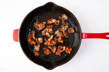 A top view of crispy fried bacon pieces in a black skillet with a red handle, set against a white background. The skillet contains some bacon grease surrounding the cooked pieces.