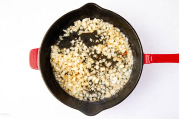 Diced onions cooking in a cast iron skillet with a red handle on a white background. The onions are starting to brown, suggesting they are being sautéed.