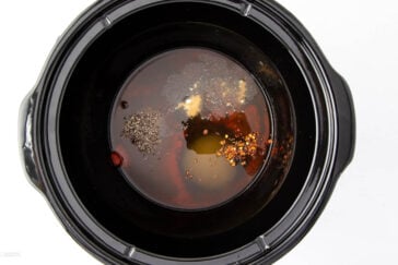 A black slow cooker with various ingredients inside, including crushed red pepper, ground black pepper, garlic powder, and liquid seasonings. The image is taken from above, showing the distinct separation of the spices and liquids.