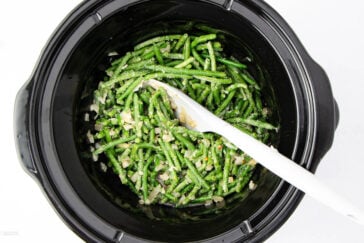 A black slow cooker filled with cooked green beans mixed with chopped onions, garlic, and seasonings. A white serving spoon is placed in the cooker, ready for serving.