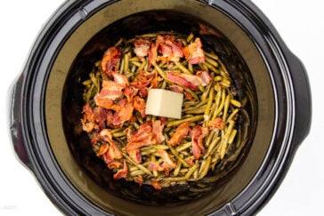 A slow cooker filled with cooked green beans, topped with crispy bacon pieces and a square of butter. The dish appears ready to serve.
