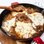 A cast iron skillet filled with cheesy French onion smothered pork chops, with a wooden spoon drizzling sauce over the top. Slices of onion are visible in the sauce, and the dish is set on a wooden board next to a striped cloth.