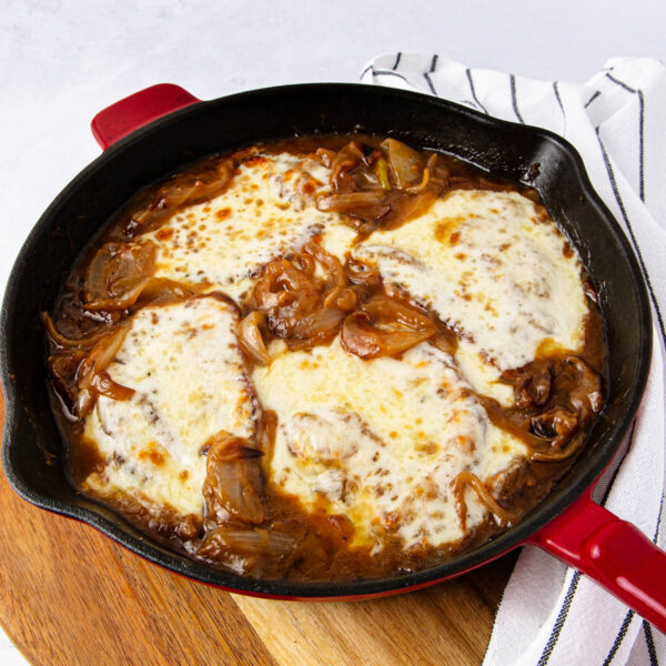 A cast iron skillet filled with french onion pork chops, smothered in melted cheese and caramelized onions, sits on a wooden board next to a white and black striped kitchen towel.