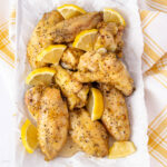 A tray of seasoned chicken wings garnished with lemon wedges is placed on parchment paper. The wings are lightly glazed and sprinkled with black pepper, surrounded by a yellow and white checkered cloth.