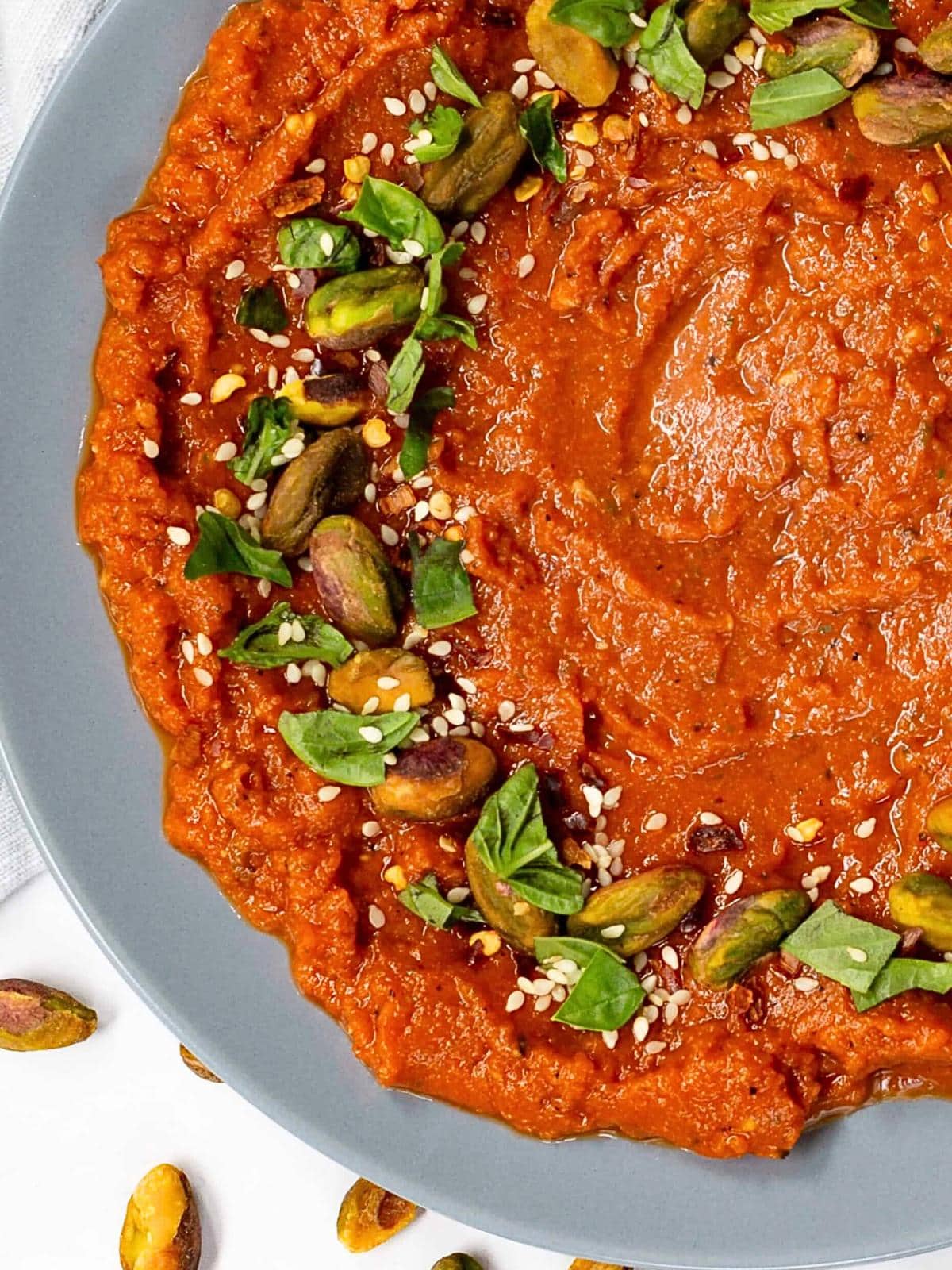 A plate of bright red muhammara dip, garnished with green pistachios, sesame seeds, and fresh basil leaves. Scattered pistachios add a delightful crunch, reminiscent of a canned cowboy candy recipe with its unique blend of sweet and spicy notes.