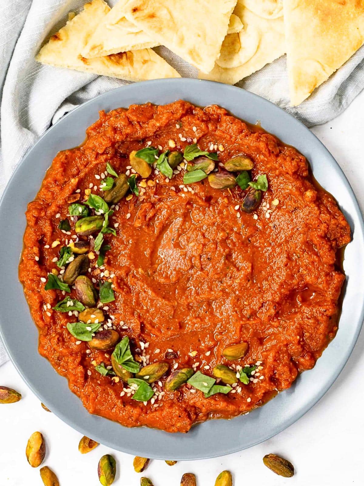 A plate of red Muhammara dip garnished with green herbs, pistachios, and sesame seeds. Pieces of flatbread sit at the top of the image, reminiscent of a homemade cowboy candy recipe, with some pistachios scattered on the light surface around the plate.