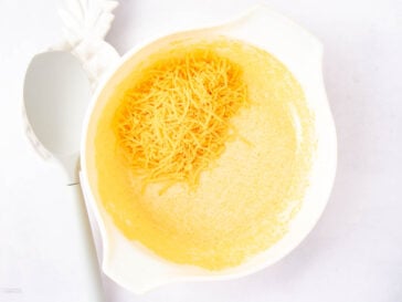 A white mixing bowl containing grated cheese on top of a light yellow batter. A white spoon rests beside the bowl. The background is a plain white surface.