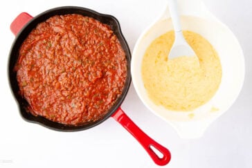 A skillet filled with rich tomato sauce is on the left, and beside it, a white bowl containing creamy polenta with a spoon rests on the right. Both dishes are placed on a white surface.