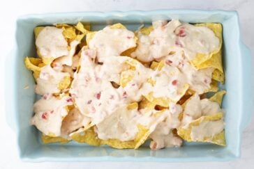 A baking dish filled with tortilla chips topped with a creamy, chunky cheese sauce containing pieces of tomato.
