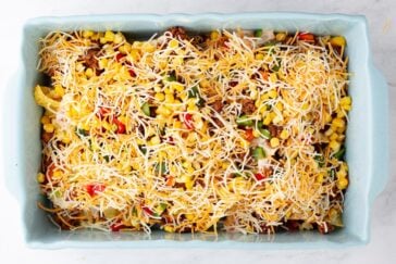 A casserole dish filled with layers of shredded cheese, corn, ground meat, and chopped red and green bell peppers, ready to be baked.
