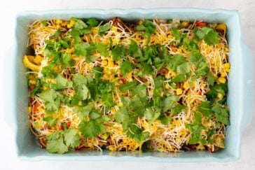 A light blue baking dish filled with a layered casserole topped with shredded cheese, corn, red bell peppers, and fresh cilantro leaves. The ingredients are colorful and evenly spread.