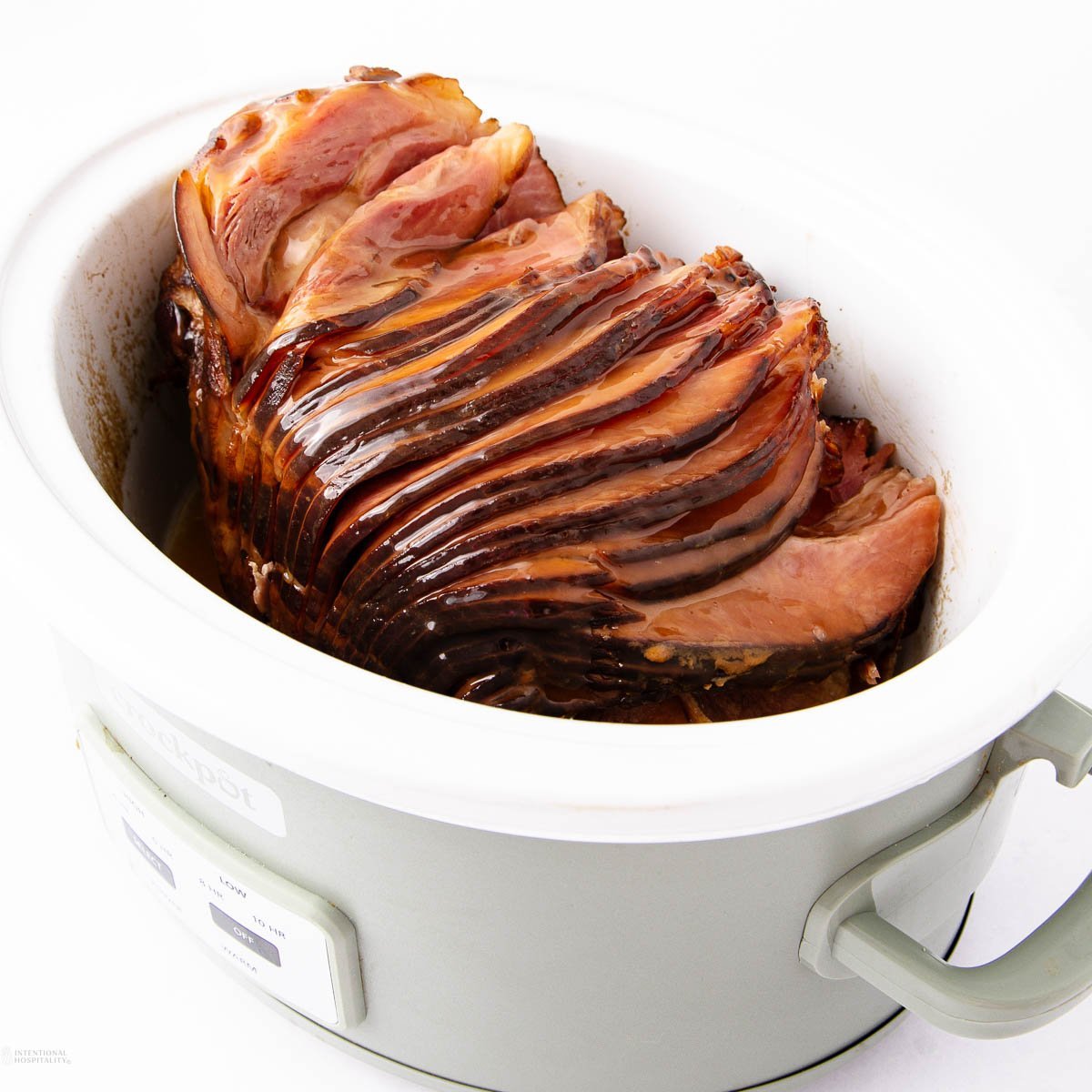 A cooked spiral ham rests in a white slow cooker. The ham is glazed and shiny, with the slices slightly separated, revealing juicy layers. The slow cooker is on a white surface, featuring control buttons on the front.