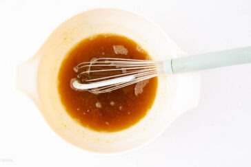A white mixing bowl filled with a brown liquid, likely a sauce or marinade, with a whisk resting inside. The bowl is placed on a white background.