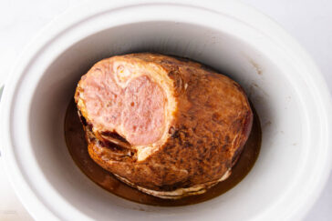 A cooked ham placed inside a white slow cooker, with its surface browned and juices collecting at the bottom. The ham appears sliced from the top portion, revealing its pink interior.