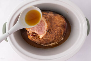 A cooked ham rests in a white slow cooker with a ladle pouring juice over it. The ham is browned and surrounded by liquid. The slow cooker has light green handles.