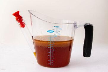 A transparent gravy separator filled with brown liquid features a black handle and a red spout stopper. It has measurement markings on the side and is placed on a white surface.