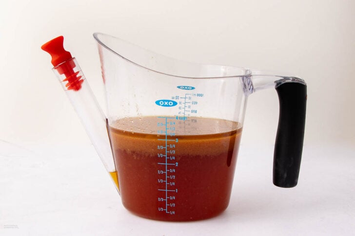 A transparent gravy separator filled with brown liquid features a black handle and a red spout stopper. It has measurement markings on the side and is placed on a white surface.