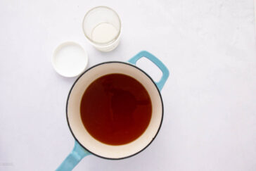 A blue-handled pot containing brown liquid sits on a white surface. Next to it, there's a small open jar with a white lid, partially filled with a light liquid.