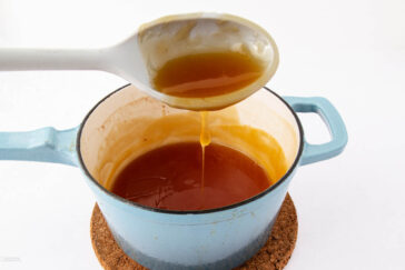 A blue pot filled with caramel sauce sits on a cork mat. A spoon is held above the pot, with caramel sauce dripping from it back into the pot, against a plain white background.