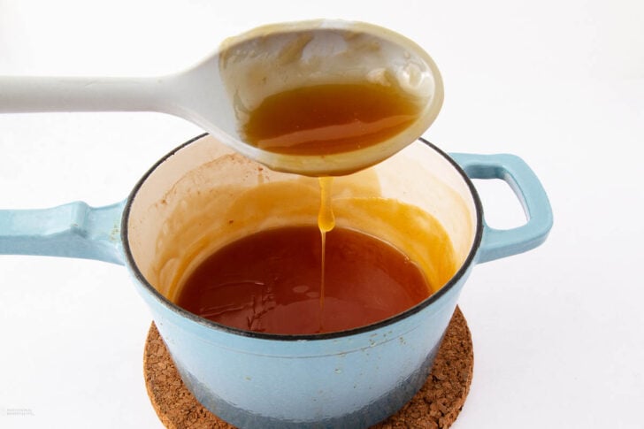 A blue pot filled with caramel sauce sits on a cork mat. A spoon is held above the pot, with caramel sauce dripping from it back into the pot, against a plain white background.