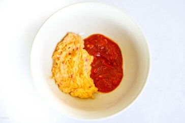 A white bowl containing a portion of creamy peanut butter soup on one side and a serving of red tomato sauce on the other, set against a plain background.