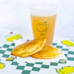 An iced drink in a "Masters 2025" plastic cup and two golden-brown baked goods sit on a paper napkin. They are placed on a paper with the Masters logo and a green-and-white checkered pattern.