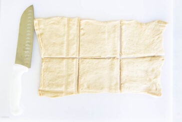 A sheet of dough is laid flat on a white surface and cut into six rectangular pieces. A large knife with a white handle rests on the left side.