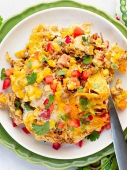 A plate of cheesy taco casserole topped with melted cheese, ground meat, corn, chopped tomatoes, and fresh cilantro, served on a white plate with a fork, placed on a green plate with a patterned napkin nearby.