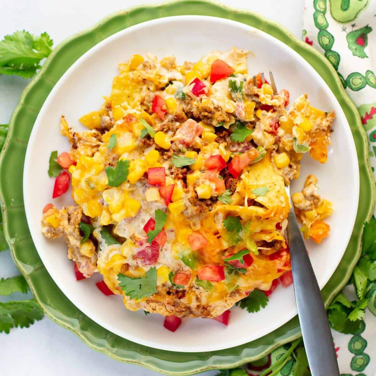 A plate of cheesy taco casserole topped with melted cheese, ground meat, corn, chopped tomatoes, and fresh cilantro, served on a white plate with a fork, placed on a green plate with a patterned napkin nearby.