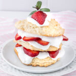 A strawberry shortcake with layers of biscuit, whipped cream, and fresh strawberry slices, topped with a dollop of cream, a strawberry half, and a sprig of mint, served on a white decorative plate.