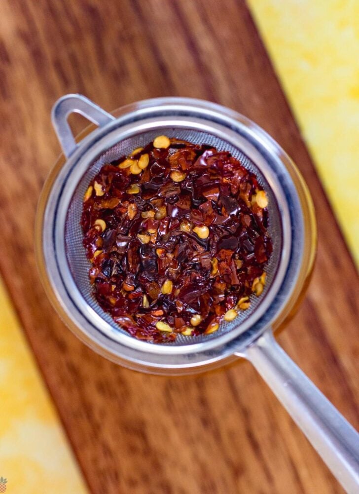 A metal strainer filled with crushed red chili flakes, perfect for adding heat to your canned cowboy candy recipe, rests on a wooden and yellow surface.