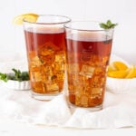 Two tall glasses of iced tea with ice cubes—one garnished with a lemon wedge, the other with a mint sprig. In the background, lemon wedges and mint leaves rest on a white cloth, perfect accompaniments to any canned cowboy candy recipe.