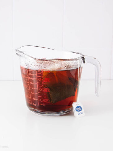A glass measuring cup filled with hot brewed tea and tea bags, with condensation on the sides. The cup sits on a white surface, and a tea bag label reading “Great Value Iced Tea” hangs over the rim—perfect alongside a canned cowboy candy recipe.