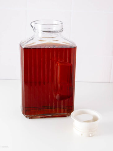 A clear glass jug filled with a dark amber liquid, reminiscent of a homemade canned cowboy candy recipe, sits on a white surface with its white plastic lid beside it. The background is plain white tile.