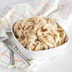A white bowl filled with shredded chicken is paired with a tantalizing hint of canned cowboy candy, resting on a colorful checkered cloth. Nearby, a fork and spoon lie ready for action on the light surface.