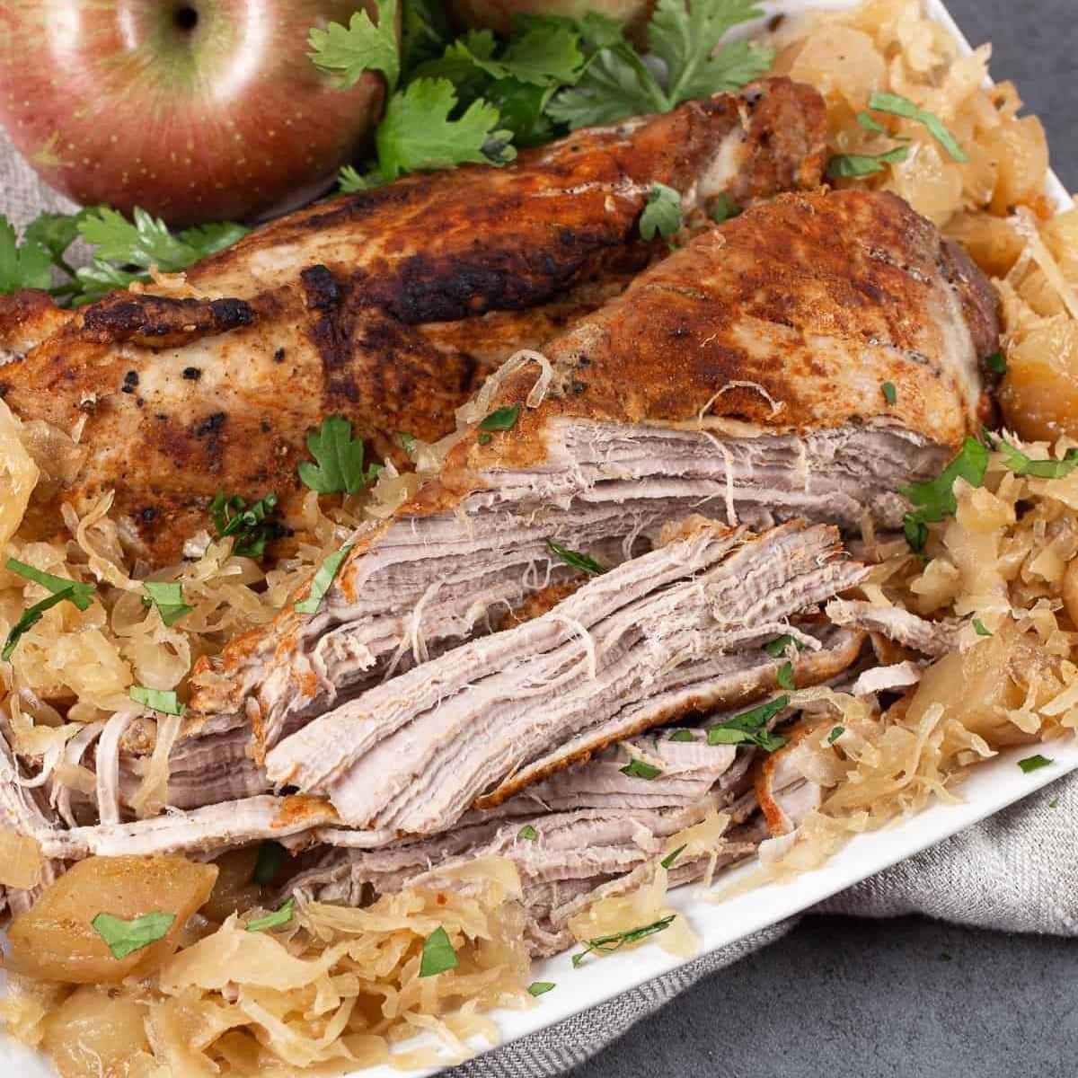 Close-up of a sliced, seasoned pork roast with sauerkraut and apple slices on a white rectangular platter. Fresh cilantro is sprinkled on top for garnish. An apple and cilantro leaves sit beside the dish, adding a hint of zest reminiscent of a spicy canned cowboy candy recipe.
