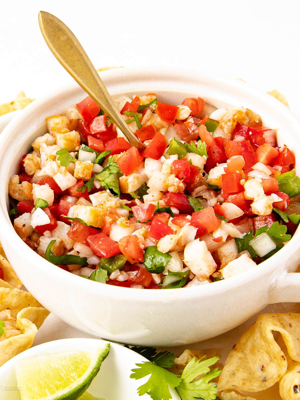 A white bowl filled with fresh ceviche made of chopped shrimp, tomatoes, onions, and peppers, garnished with cilantro, with a spoon in the bowl. Tortilla chips, lime wedges, and cilantro surround the bowl.