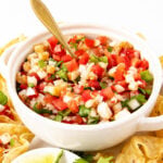 A white bowl filled with fresh ceviche made of diced tomatoes, onions, peppers, and herbs, with a gold spoon inside. Tortilla chips and a wedge of lime are visible around the bowl.