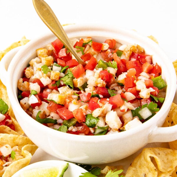 A white bowl filled with fresh ceviche made of diced tomatoes, onions, peppers, and herbs, with a gold spoon inside. Tortilla chips and a wedge of lime are visible around the bowl.