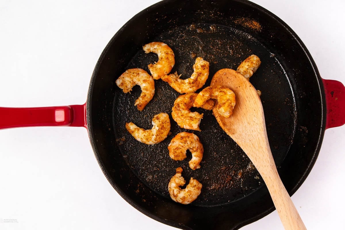 Cooked, seasoned shrimp in a black skillet with a red handle, being stirred with a wooden spoon, set against a white background.