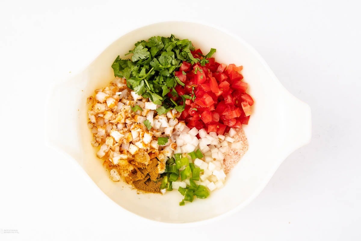 A white bowl containing chopped cilantro, tomatoes, onions, green peppers, and hard-boiled eggs, along with a mix of seasonings, on a white background.