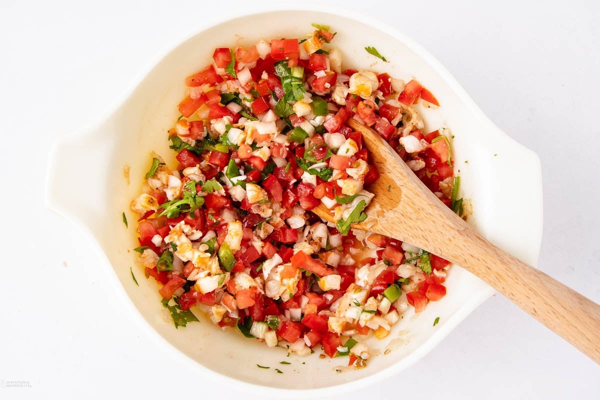 A white bowl filled with freshly chopped vegetables, including tomatoes, onions, cilantro, and peppers, being mixed with a wooden spoon. The mixture resembles a salsa or fresh salad.