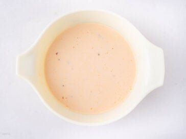A white bowl filled with a pale orange creamy liquid, possibly a sauce or soup, placed on a white surface.