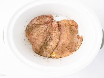 Two raw, seasoned chicken breasts placed inside a white slow cooker, viewed from above against a white background.
