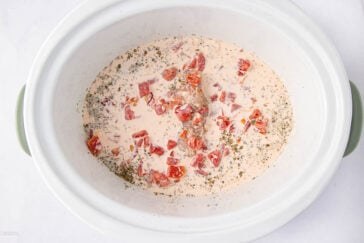 A slow cooker filled with raw chicken breasts, diced tomatoes, cream, and herbs, ready to be cooked.