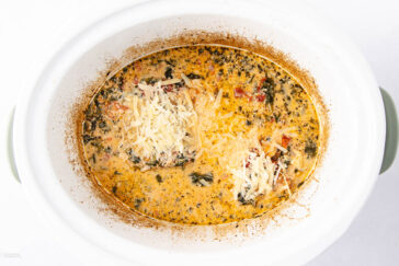 Creamy soup with herbs, tomatoes, and spinach topped with shredded cheese in a white slow cooker.