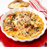 A white dish filled with creamy tomato chicken, fusilli pasta, spinach, and topped with shredded cheese; a bowl of grated cheese and a red striped napkin are nearby on a red plate.